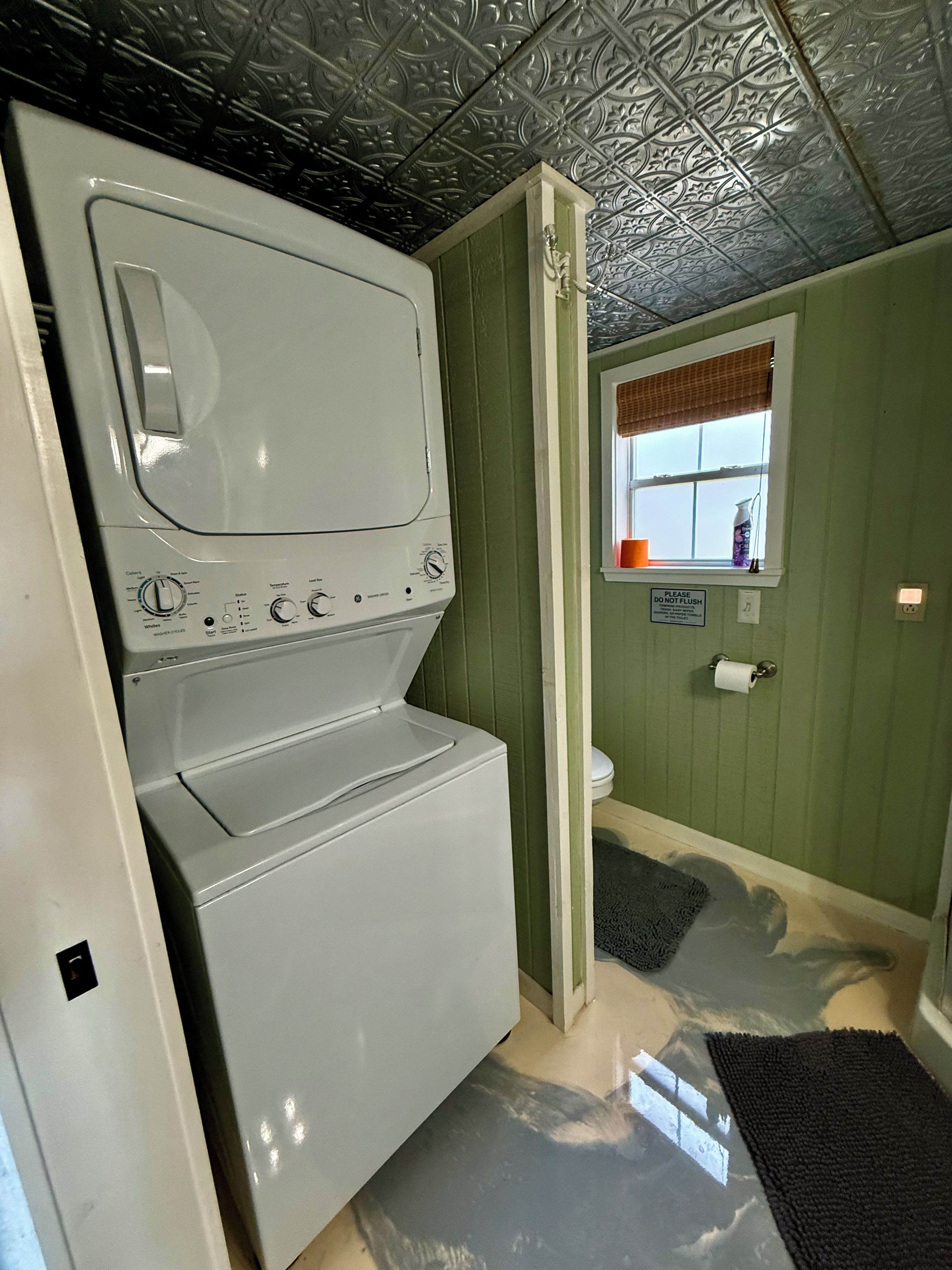 Une buanderie équipée d’un lave-linge et d’un sèche-linge superposés, d’une fenêtre offrant une belle vue et d’un panneau indiquant : « Veuillez ne pas jeter de déchets dans les toilettes ».