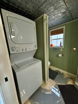 A laundry room with a stacked washer and dryer, a window with a view, and a 'Please do not flush' sign. - Entire guesthouse in New Braunfels, Texas (New Braunfels)
