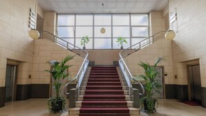 A grand staircase with marble steps and a red carpet, flanked by potted plants and metal railings, leading to a high ceiling with large windows. - Prestige Apartment Via Roma by Wonderful Italy (Turin)