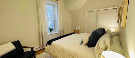 A bedroom with a bed, a desk, a chair, a ceiling fan, a window with curtains, and a bedside lamp.