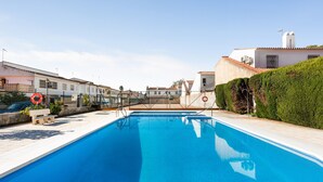 A swimming pool with a clear blue water surface, surrounded by a white building with a tiled roof, and a green hedge on the right.