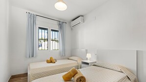 A hotel room with two beds, a window with curtains, an air conditioning unit, a bedside table with a lamp, and a wooden floor.