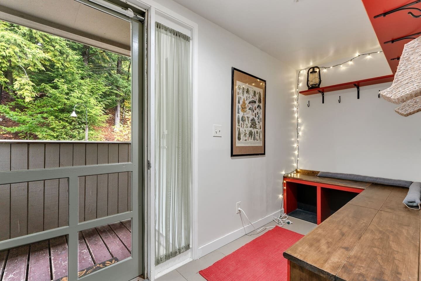 Une pièce avec une terrasse en bois, un banc rouge, une petite table, une illustration botanique encadrée et une guirlande lumineuse.