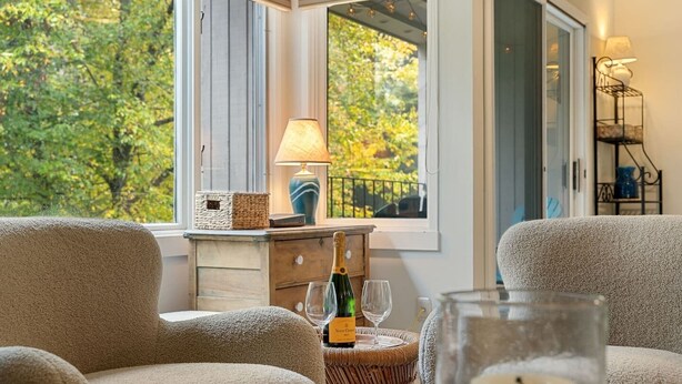 Une pièce chaleureuse avec vue sur les arbres, une petite table d’appoint en osier sur laquelle se trouvent une bouteille et des verres, et une lampe sur une table de chevet.