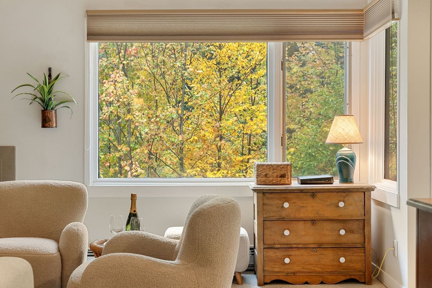 Una habitación con un ventanal grande, un aparador de madera, una lámpara, una botella y una planta.