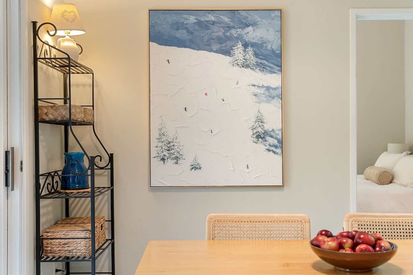 Un comedor con una mesa de madera, sillas de mimbre y una estantería metálica con cestas y un jarrón azul.