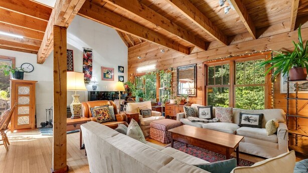 A cozy, rustic living room with wooden beams, a fireplace, and a large window overlooking greenery.