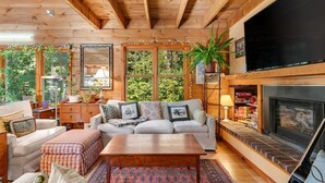 A cozy living room with a wooden ceiling, a fireplace, a television, and a large window overlooking greenery.
