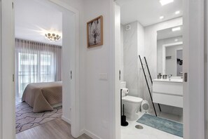 Een moderne hotelkamer met een bed, een badkamer met toilet, wastafel en spiegel, en een gang die naar een andere kamer leidt.