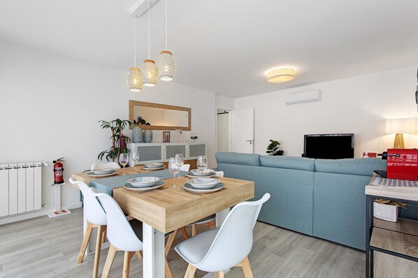 A modern living room with a dining area, a blue sofa, a TV, and a wooden dining table with white chairs.