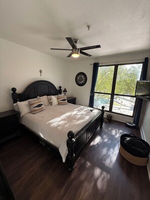 Un dormitorio con cama, ventilador de techo, reloj, ventana con cortinas y una planta pequeña.