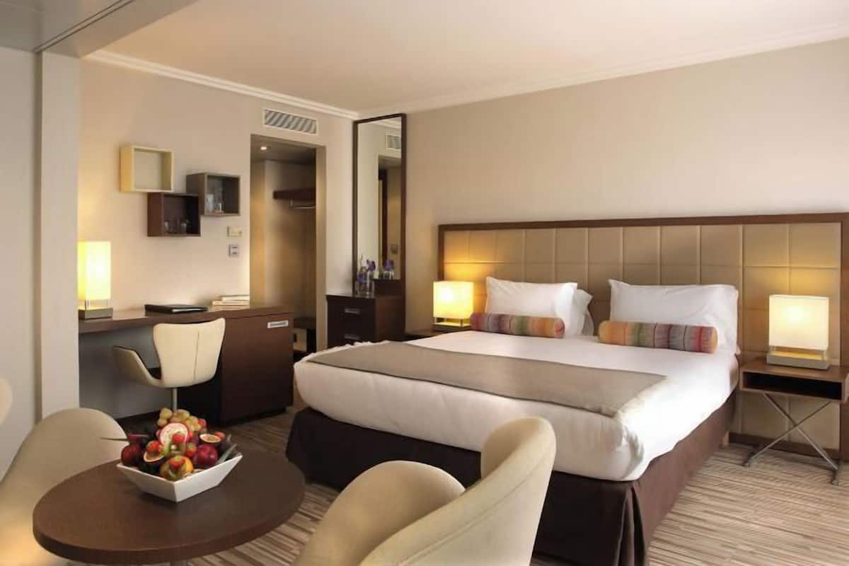 Quarto de hotel com cama, mesas de cabeceira, escrivaninha, cadeira, mesa redonda com frutas e espelho.