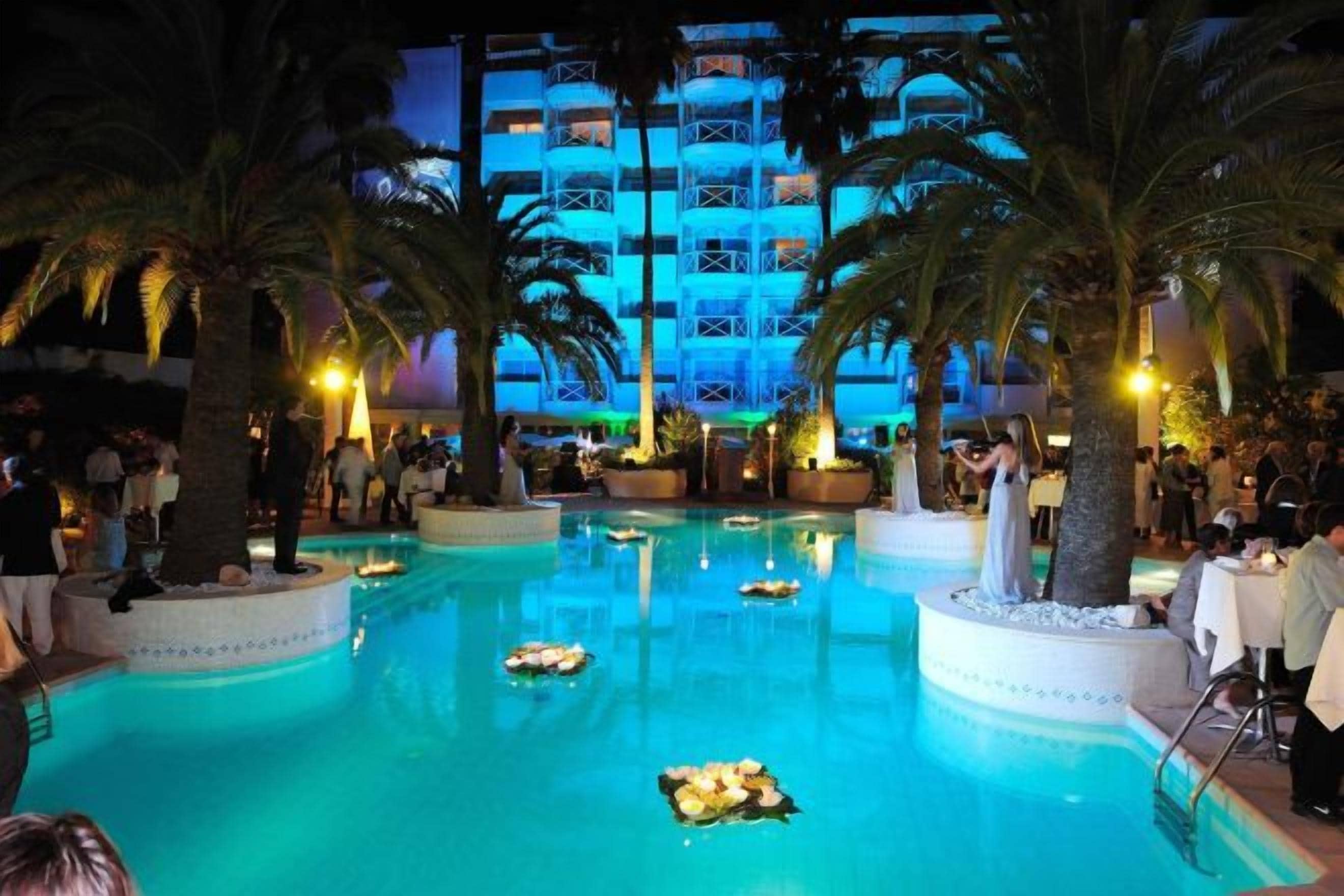 A pool area with multiple swimming pools, palm trees, and a building with balconies in the background.