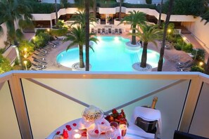 Un espace piscine avec des chaises longues et des palmiers, un coin repas avec une table et des chaises, et une vue sur un immeuble à plusieurs étages.