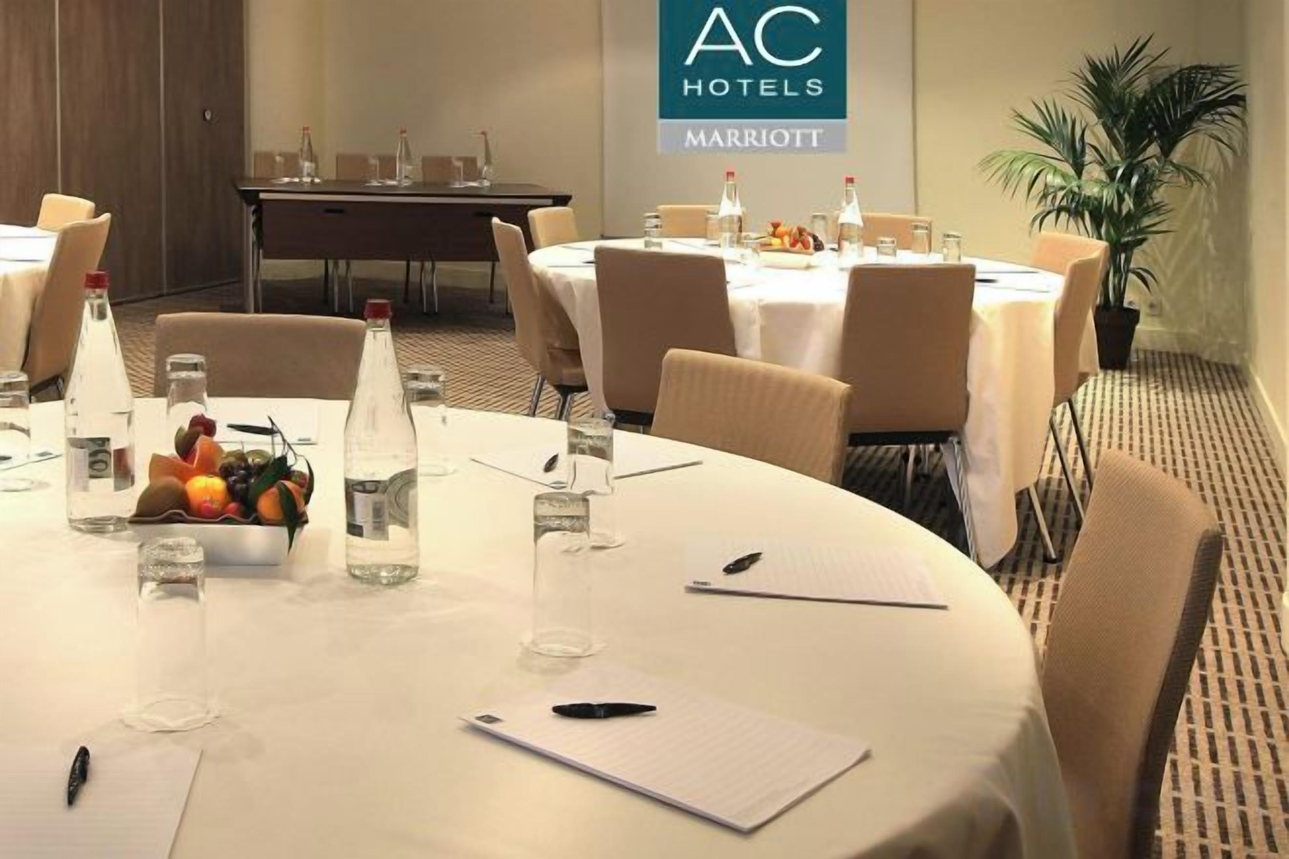 Ein Konferenzraum mit weißem Tisch, Stühlen, Wasserflaschen, einer Obstschale und einem Schild für AC Hotels Marriott.