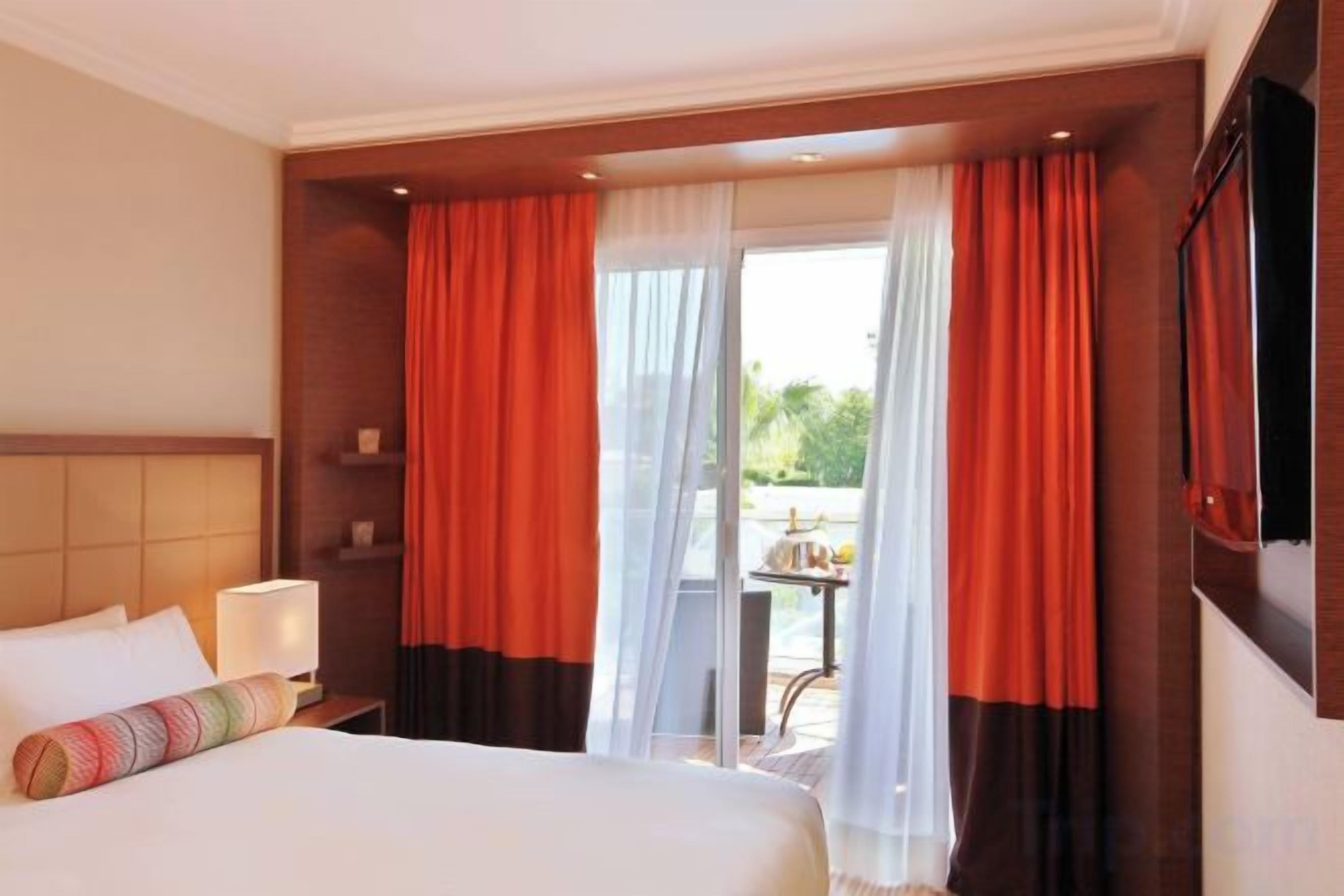 Ein Hotelzimmer mit einem Bett, einem Nachttisch, einer Lampe und Blick ins Freie durch eine Glastür.