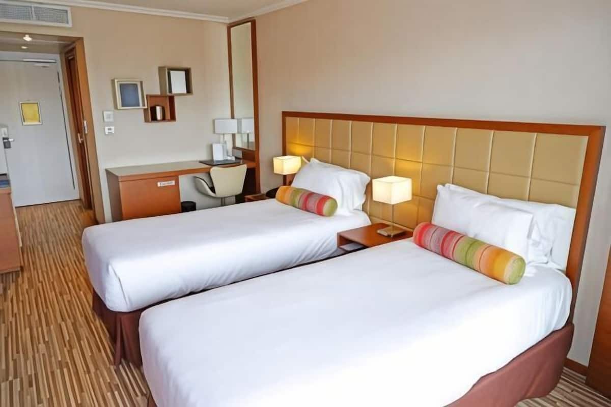 Quarto de hotel com duas camas, uma escrivaninha, uma cadeira e um espelho.