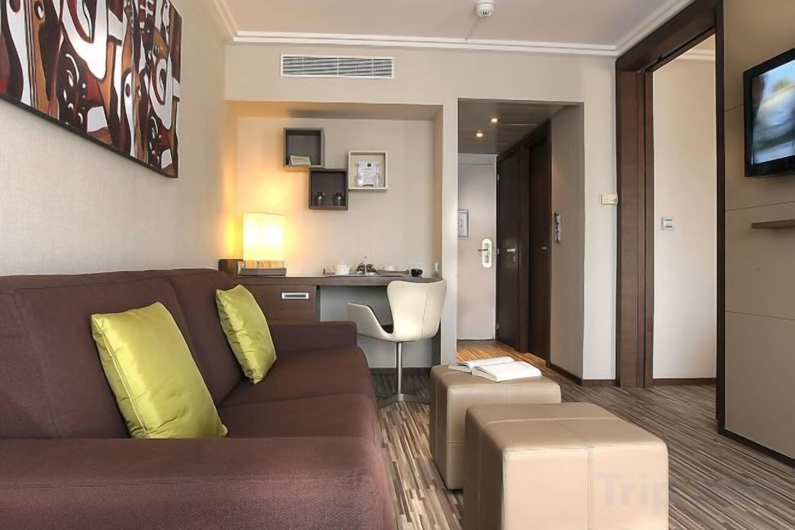 Une chambre d’hôtel avec un canapé marron, un petit bureau, une chaise et un téléviseur fixé au mur.