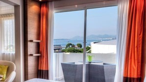A hotel room with a bed, a chair, a small table, and a balcony with a view of the sea and buildings. - AC Hotel by Marriott Ambassadeur Antibes - Juan Les Pins (Juan-les-Pins)