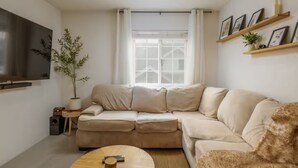 En moderne stue med en beige modulsofa, et træsofabord, et fladskærms-TV monteret på væggen, en potteplante og et vindue med hvide gardiner.