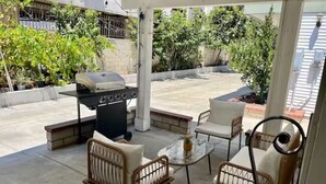 En overdækket terrasse med grill, fletstole og et glasbord.