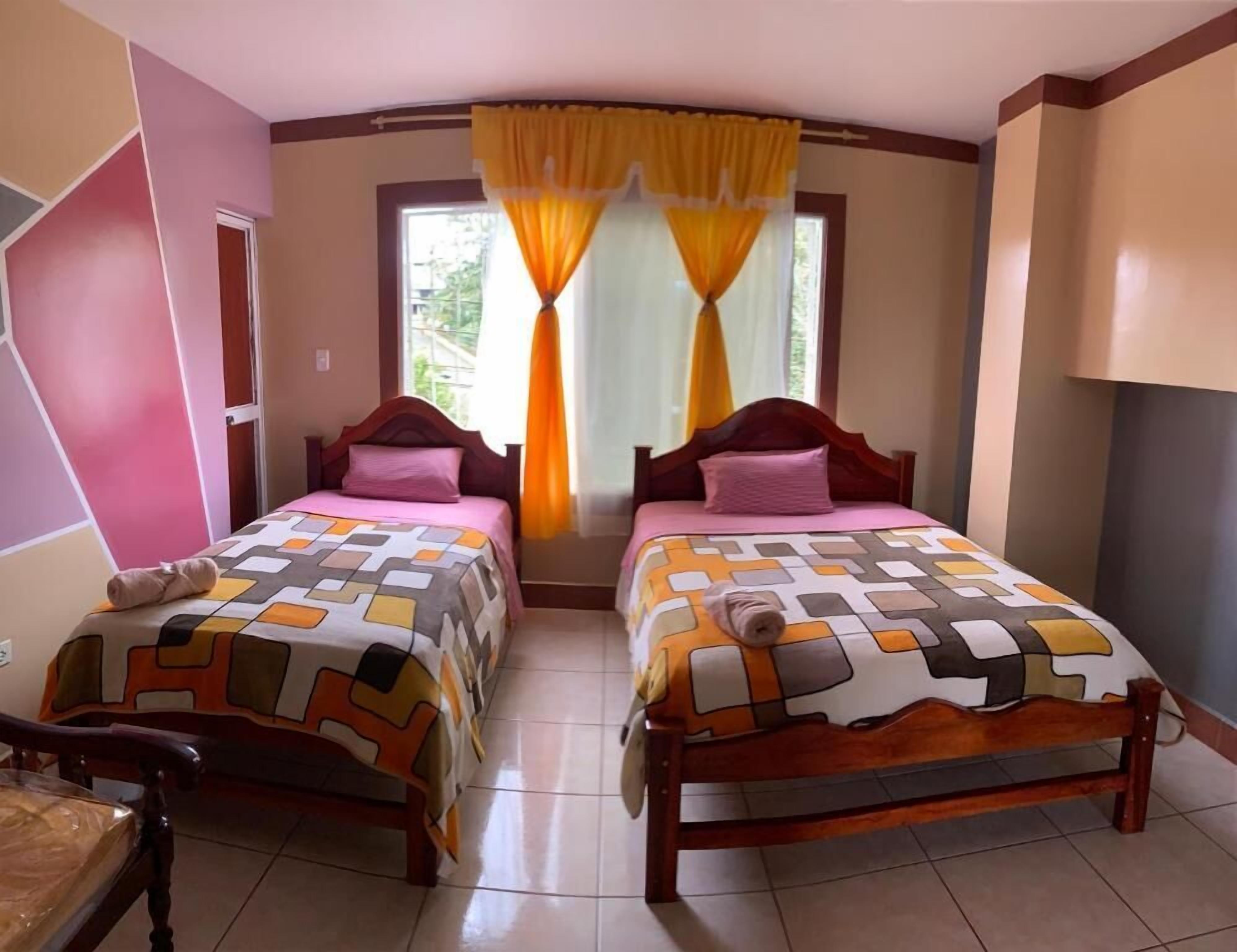 A room with two beds, each with a patterned bedspread, a wooden headboard, and a window with yellow curtains.