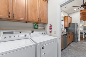 A kitchen with wooden cabinets, a Whirlpool washer, a Whirlpool dryer, a stainless steel refrigerator, and a fire extinguisher mounted on the wall.