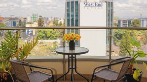 Un balcon avec deux fauteuils en osier, une petite table ronde ornée d’un vase de fleurs, et une vue sur le paysage urbain, notamment sur l’immeuble West End Towers.