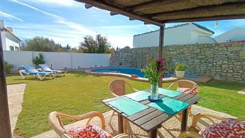 A covered outdoor area with a table and chairs, a pool, and a stone wall in the background.