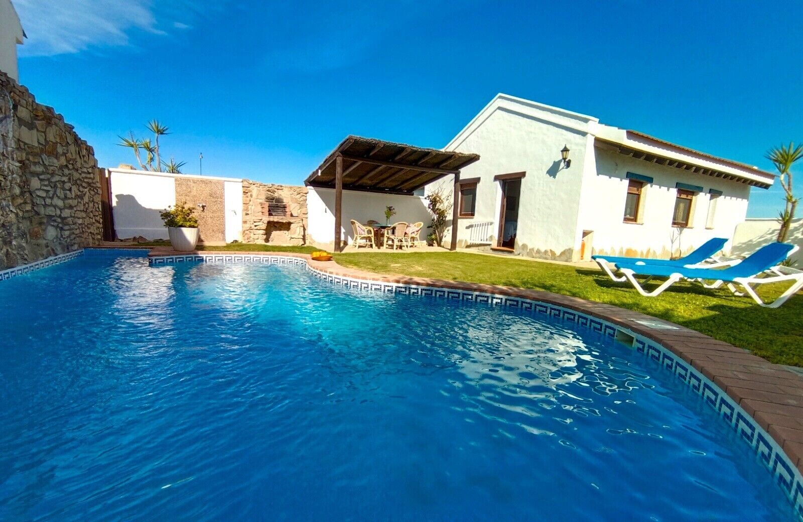 Una piscina con muro de piedra, un porche cubierto y un edificio de fachada blanca y ventanas azules.