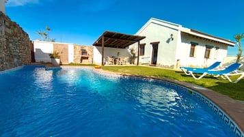 Una piscina con muro de piedra, un porche cubierto y un edificio de fachada blanca y ventanas azules.