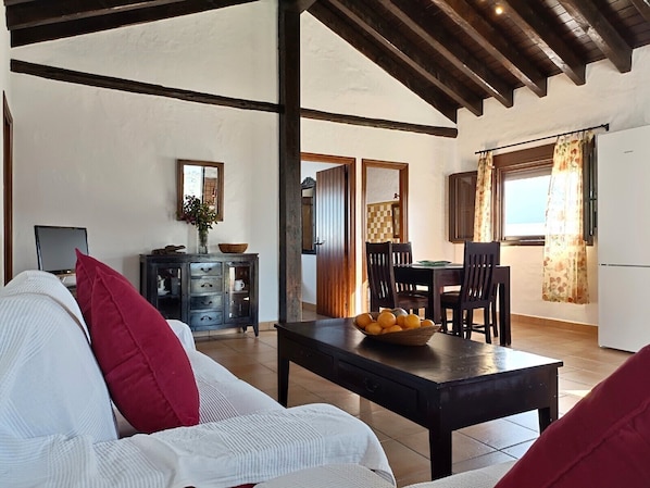 A living room with a sofa, a dining table with chairs, a TV, and a kitchen area with a window.