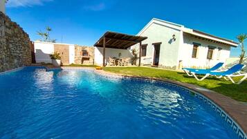 Una piscina con borde de piedra, un porche cubierto y un edificio de fachada blanca y ventanas azules.