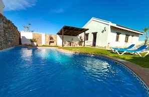 A swimming pool with a stone border, a covered patio, and a building with a white facade and blue windows.