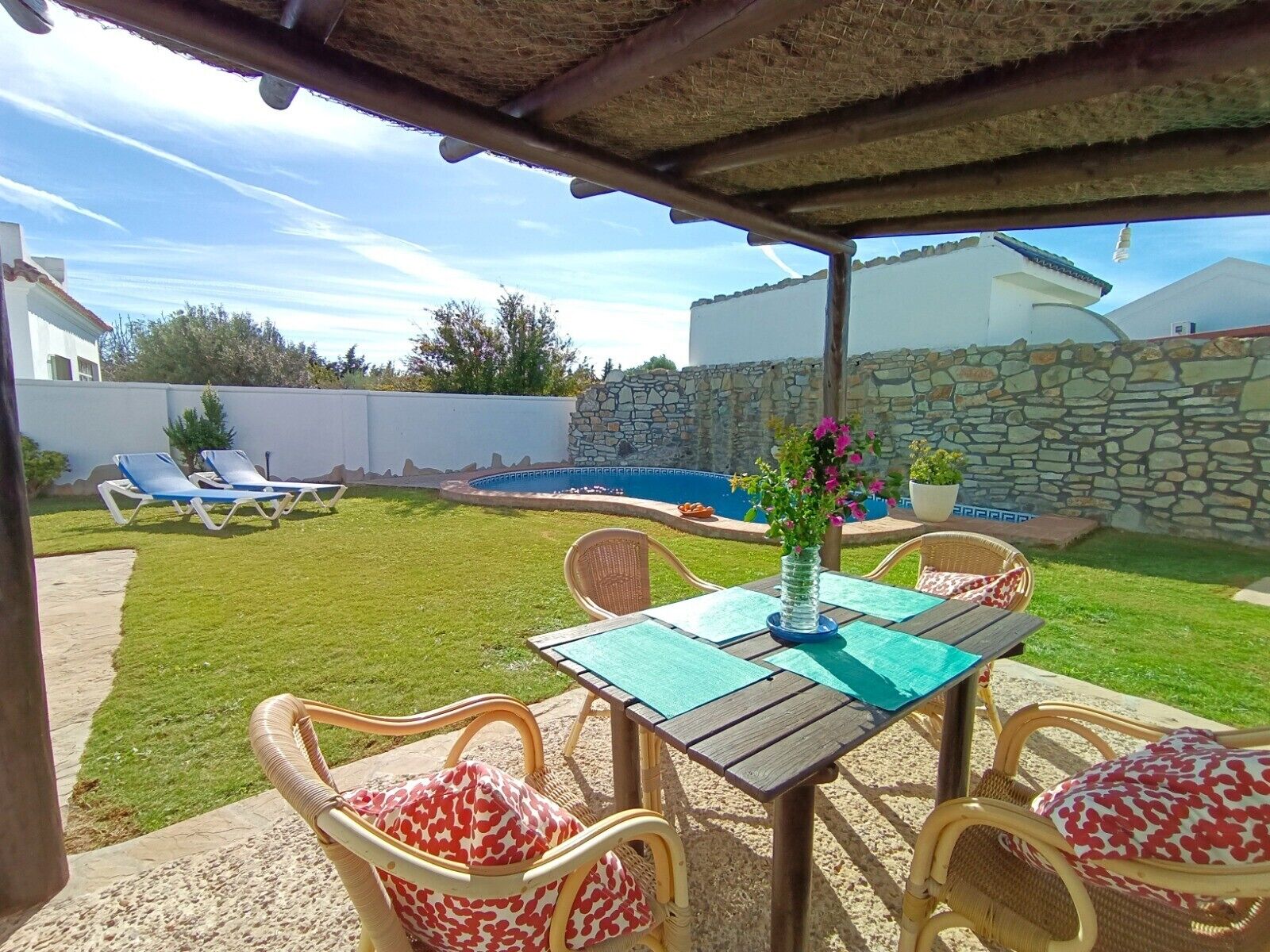 Un espacio de estar al aire libre cubierto con mesa y sillas, una piscina y un muro de piedra al fondo.