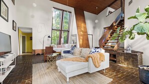 Un salon spacieux avec un canapé blanc, une table basse et une grande fenêtre donnant sur des arbres. On aperçoit un escalier avec une rampe en bois au fond.