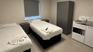 Une chambre moderne avec deux lits, une armoire, un petit coin cuisine et une fenêtre avec des stores.