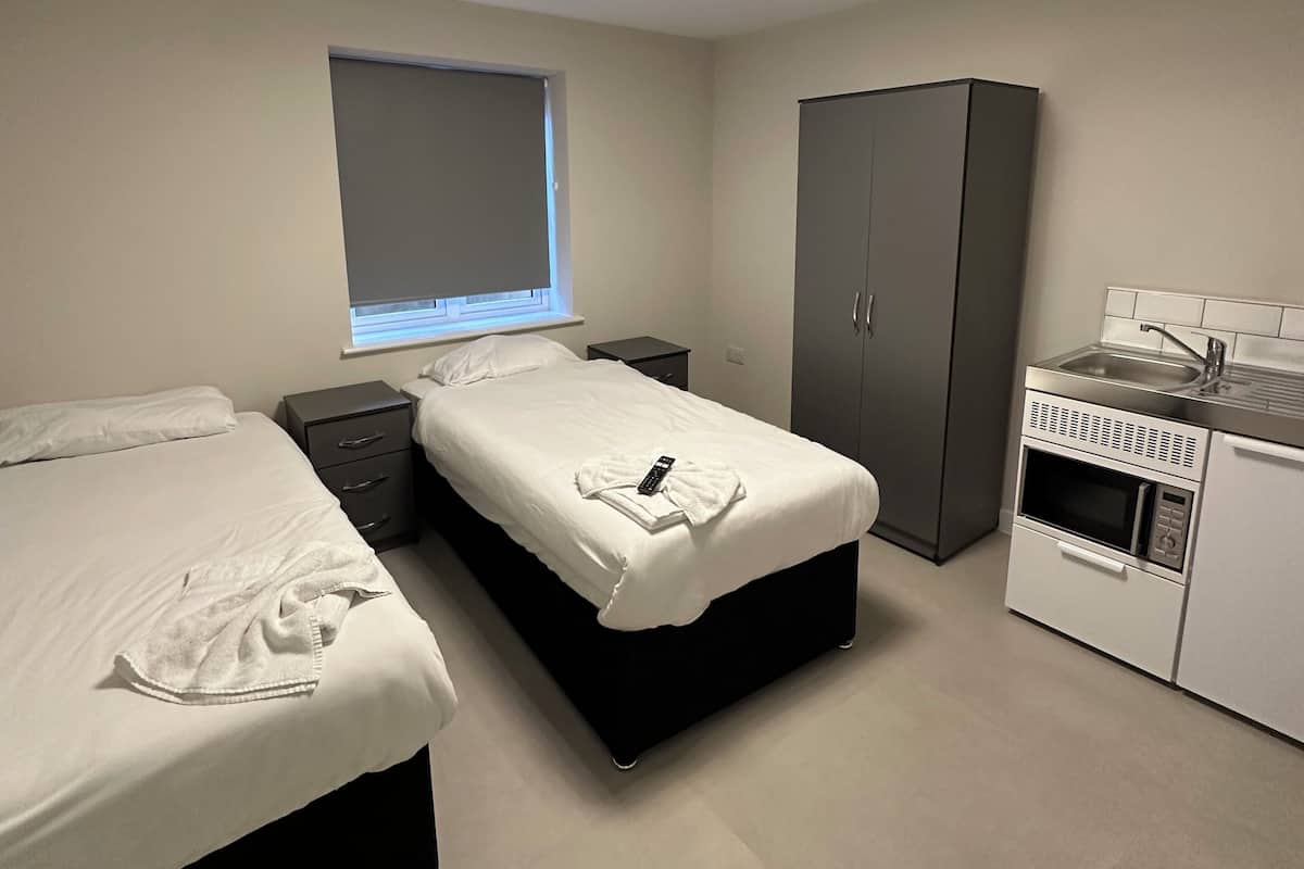 A modern bedroom with two beds, a wardrobe, a small kitchenette, and a window with blinds.