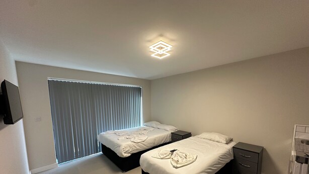 Une chambre moderne avec deux lits, une table de chevet, un téléviseur fixé au mur et un luminaire au plafond.