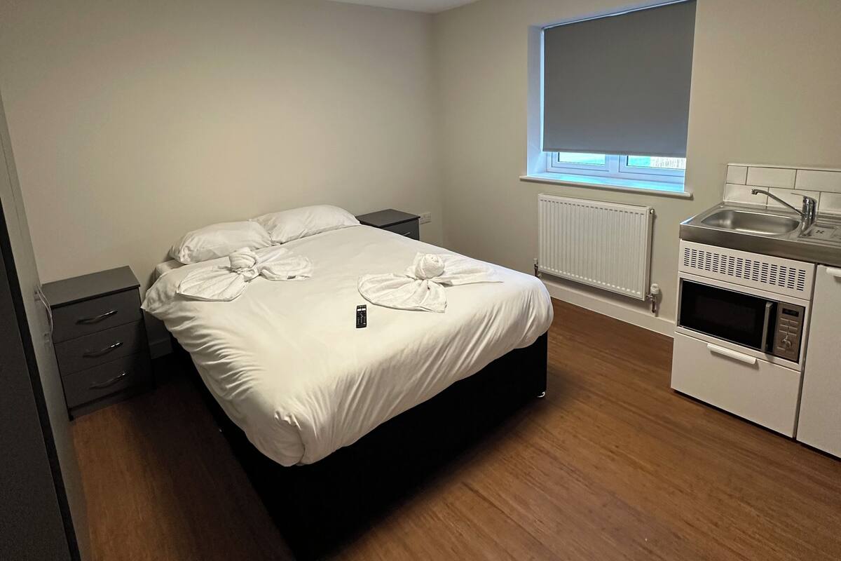 A modern bedroom with a bed, bedside tables, a built-in kitchenette, and a window with blinds.
