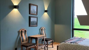 Una stanza con due sedie di legno, un tavolino e un letto con copriletto floreale. Sono presenti due luci a parete e due quadri appesi al muro.