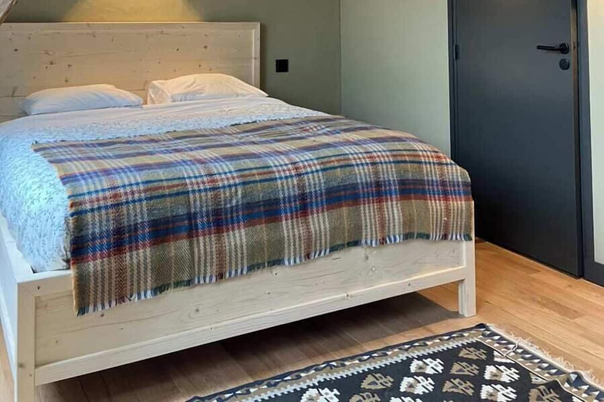 A bedroom with a wooden bed, a plaid blanket, a dark door, and a wall-mounted light fixture.