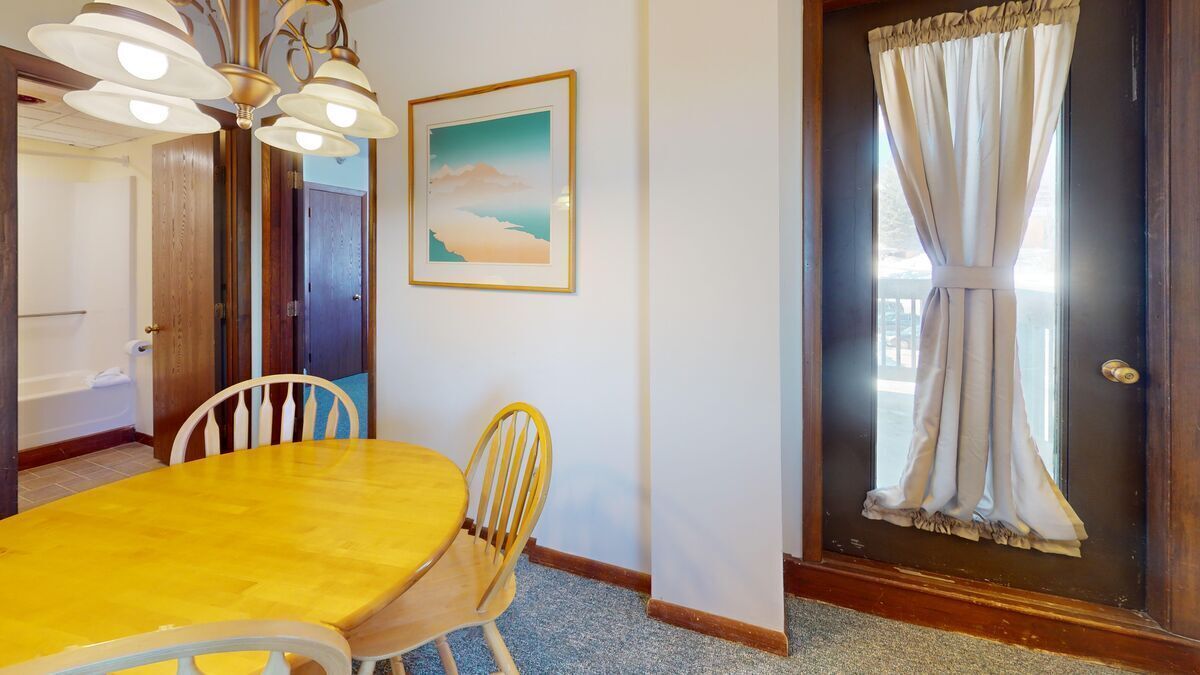 A dining area with a yellow table and white chairs, a framed picture on the wall, and a door with curtains.