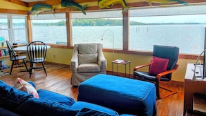 A room with a wooden floor, a blue sofa, a white armchair, and a black chair. There are two ceiling fans, a window with a view of a lake, and a shelf with books. - Water Lovers Retreat: stunning water views - dolphins for neighbors (Peletier)
