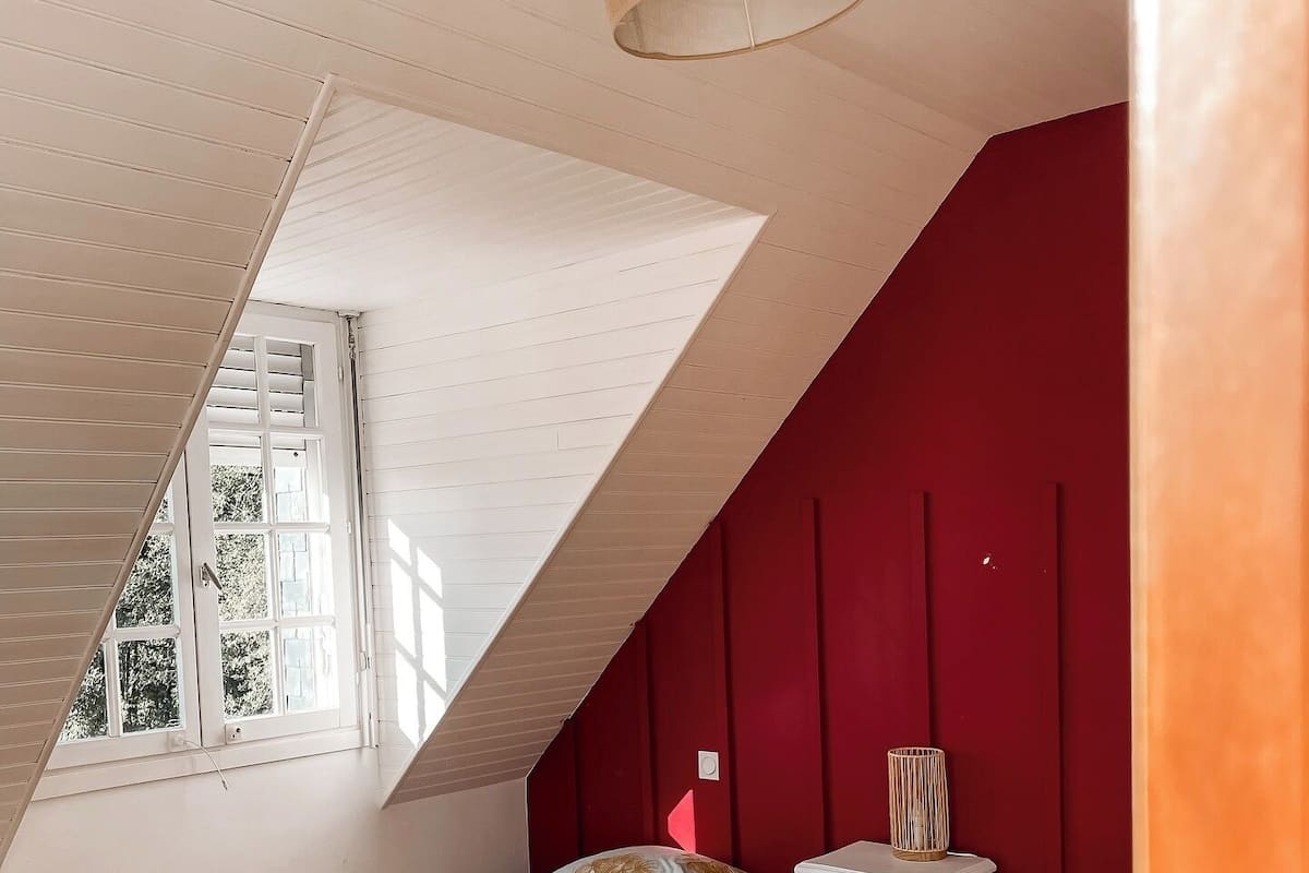 A bedroom with two beds, a window, a red wall, and a sloped ceiling.