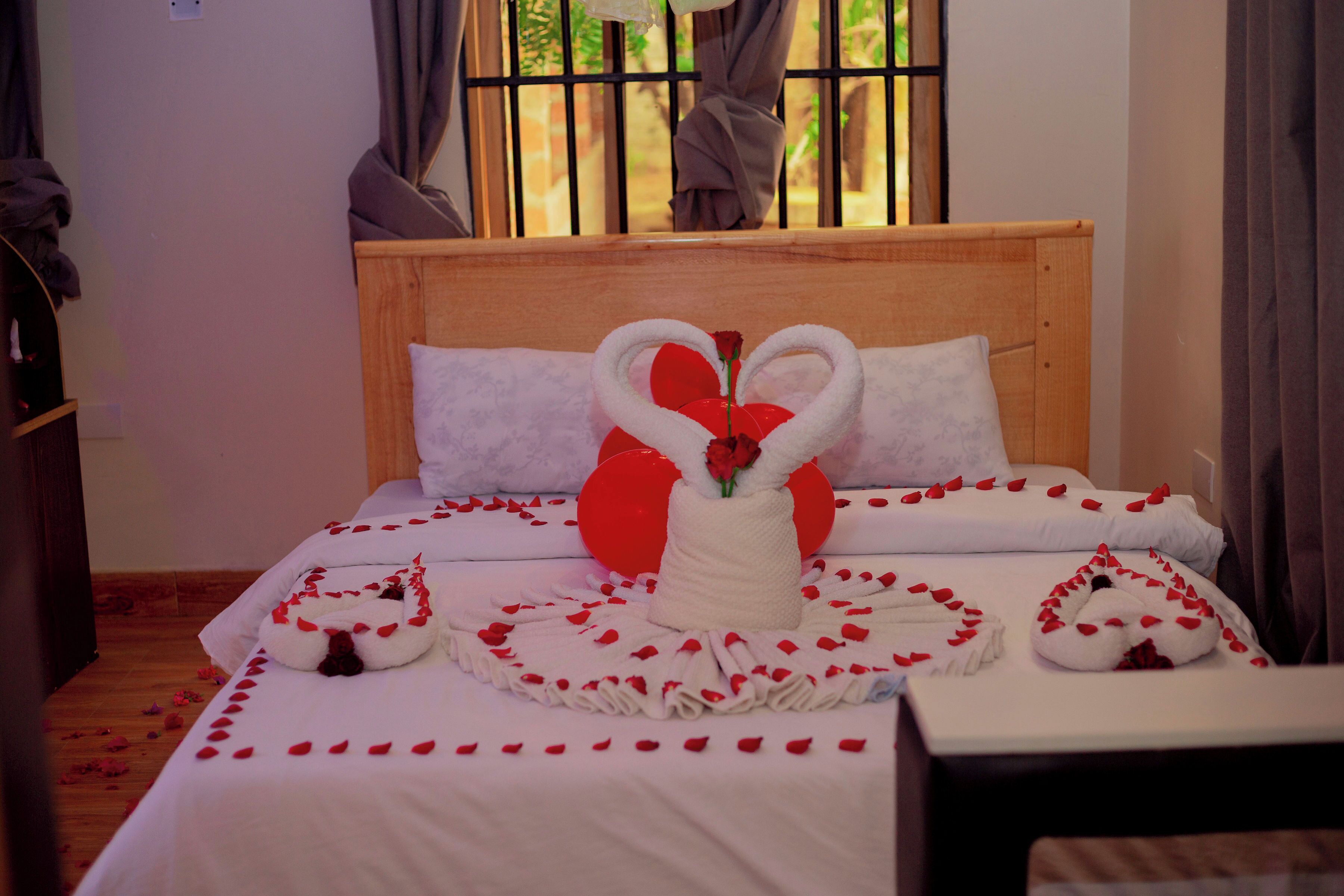 A bed with a heart-shaped towel arrangement, a vase with a rose, and red rose petals scattered on the bed.