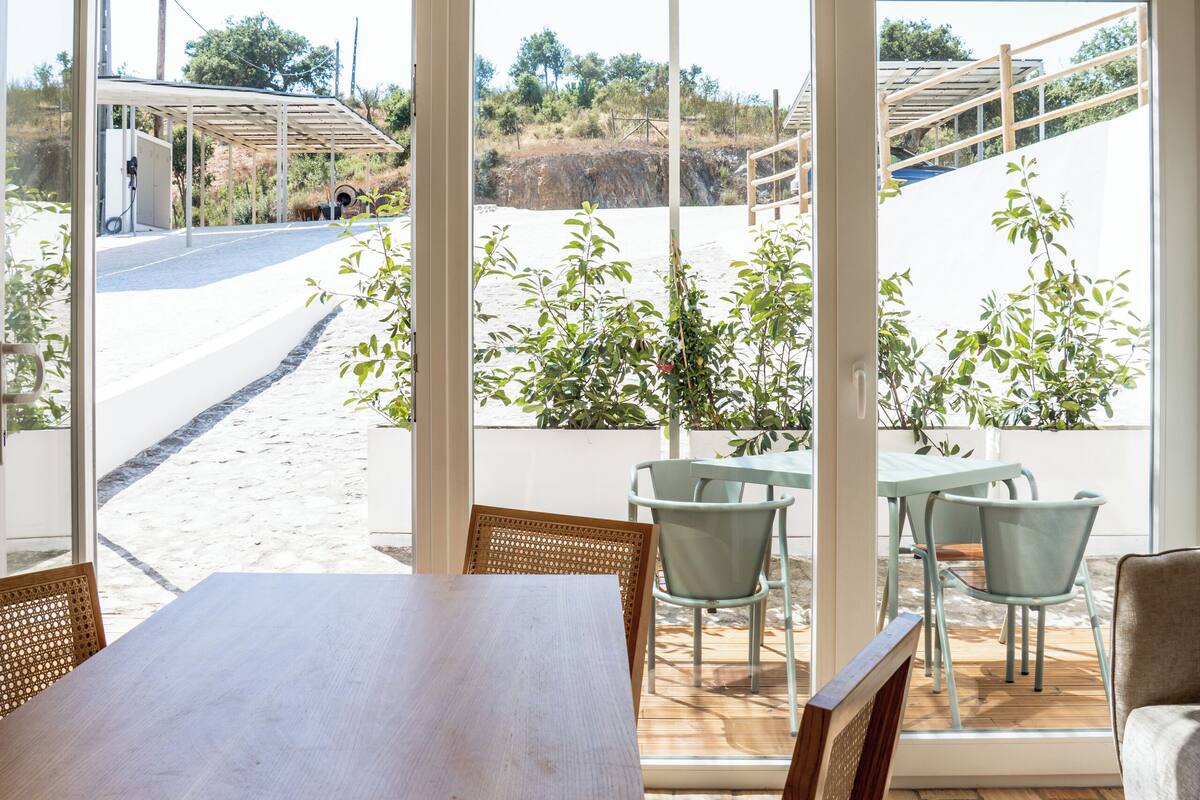 Zona de menjador amb taula i cadires de fusta, grans finestres que donen a una carretera i vegetació a l'exterior.