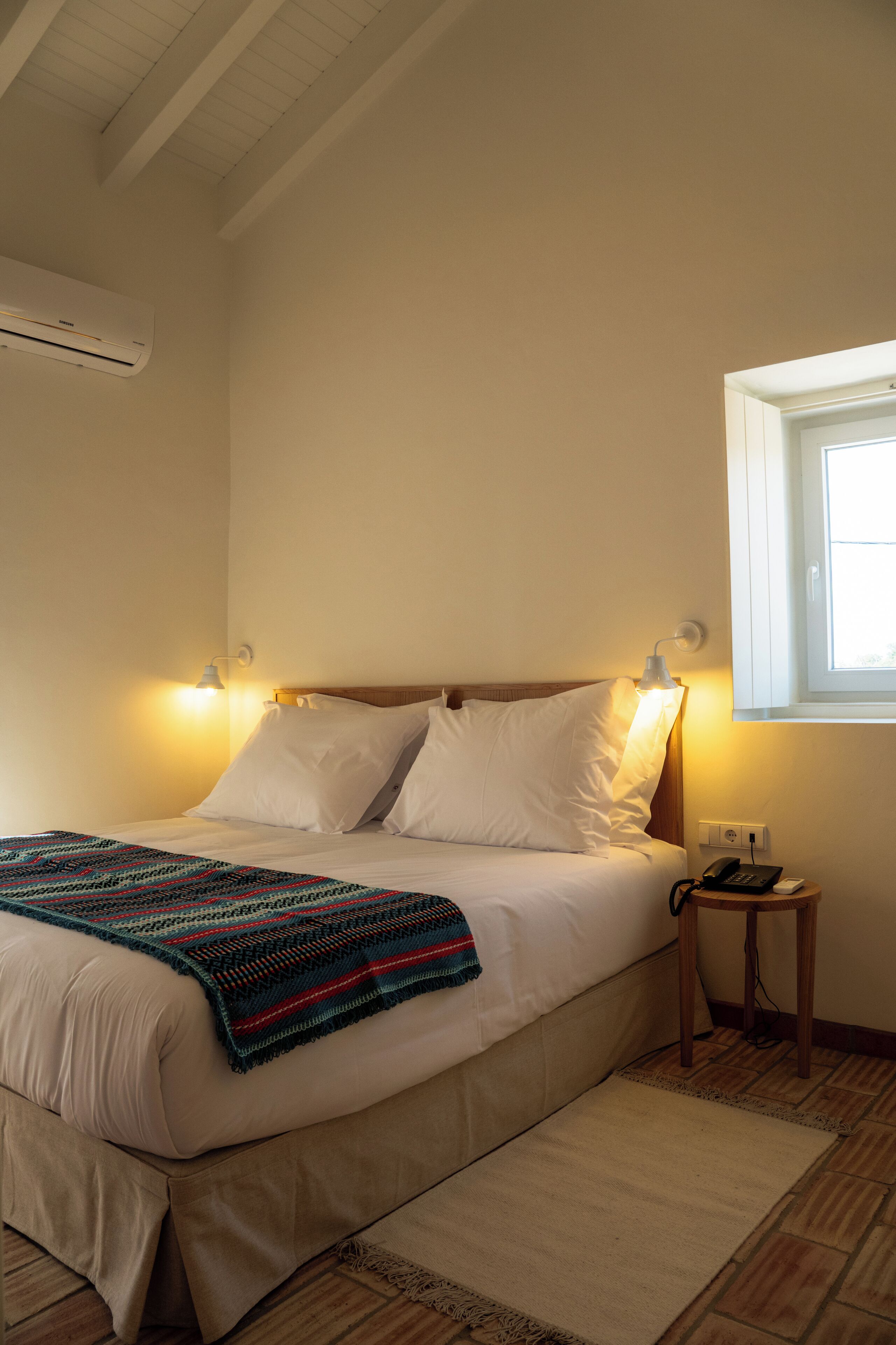 Un letto rifatto con cura, una coperta colorata, un comodino con telefono e caricabatterie, e una finestra da cui entra la luce del giorno.