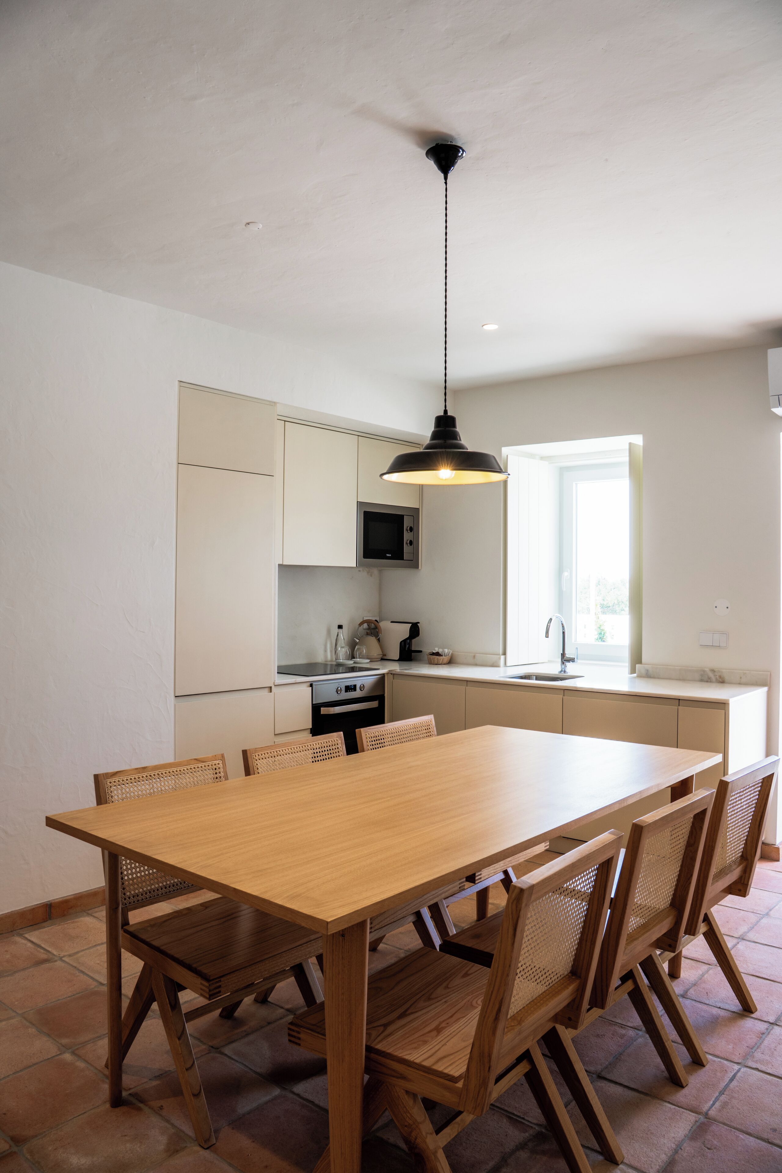 A modern kitchen with a wooden dining table and chairs, a hanging light fixture, and a window providing natural light.