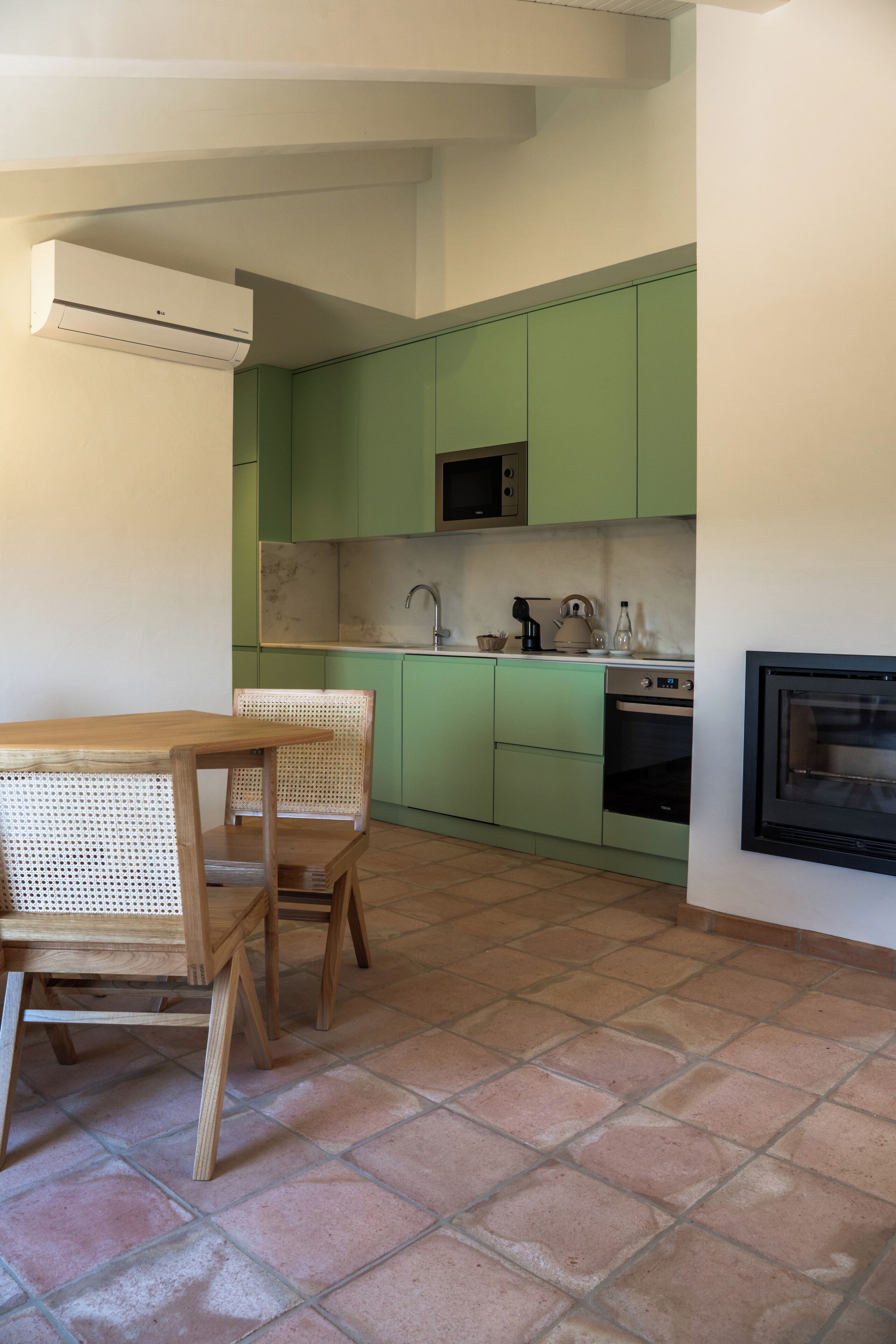 Een kleine keuken met groene kastjes, een magnetron, een oven en een eethoek met een houten tafel en rieten stoelen.